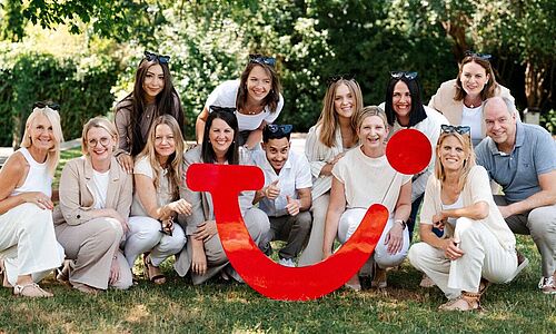 15 Menschen knien im Freien auf einer Wiese hinter einem großen roten TUI-Logo und lächeln in die Kamera. Sie sind alle hell gekleidet.