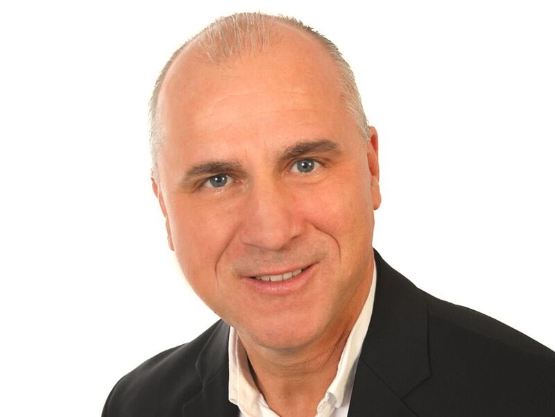 Porträt eines Mannes mittleren Alters, der in die Kamera lächelt. Er trägt ein weißes Hemd unter einer grauen Anzugjacke