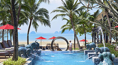 Luxuriöses Strandresort mit einem Pool, gesäumt von Palmen und bronzenen Elefantenbrunnen, die Wasser in das Becken spritzen. Im Hintergrund befinden sich rote Sonnenschirme am Sandstrand mit Blick auf das türkisfarbene Meer und ferne Inseln. Ein runder Steintorbogen rahmt den Zugang zum Strand