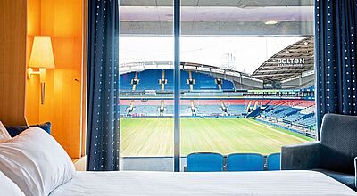 Blick in ein Hotelzimmer, hinter einem mit weißer Bettwäsche bezogenen Bett, über dem eine Nachttischlampe angeknipst ist, sieht man durch ein Fenster in den Innenraum eines Stadions