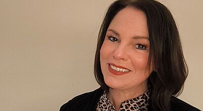 Porträt einer lächelnden Frau mit schulterlangen dunklen Haaren. Sie trägt eine schwarz gemusterte Bluse mit einem schwarzen Jackett und hat ein freundliches Lächeln. Der Hintergrund ist neutral beige