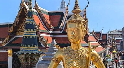 Kurzzeitig für Touristen geschlossen: der Wat Phra Kaeo in Bangkok Goldene Statue einer himmlischen Wächterfigur im Tempel Wat Phra Kaeo in Bangkok, im Hintergrund sind reich verzierte Tempeldächer und Besucher auf dem Tempelgelände zu sehen.