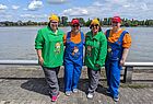 Vier Frauen stehen lächelnd nebeneinander am Ufer eines Flusses bei sonnigem Wetter. Zwei von ihnen tragen grüne Oberteile mit einer Zeichentrickfigur darauf, graue Hosen und gelbe Zipfelmützen. Die anderen beiden sind in bunten Latzhosen-Outfits gekleidet, mit orangen Oberteilen und blauen Hosen, ebenfalls verziert mit einer Comicfigur. Beide tragen rote Kappen mit „Emirates“-Aufschrift. Im Hintergrund sind Wasser, Bäume und Gebäude auf der gegenüberliegenden Flussseite zu sehen. Die Stimmung wirkt fröhlich und entspannt.