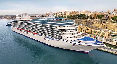 Das Kreuzfahrtschiff Vista kann unter anderem in Kopenhagen, Warnemünde und Barcelona angeschaut werden Ein hochaufragendes Kreuzfahrtschiff liegt an einem Kai, dahinter die nicht so hohen Gebäude einer Stadt.