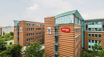 Dertour-Zentrale in Frankfurt am Main: Der Konzern gibt sich eine neue Mangement-Struktur.  Mehrstöckiges Bürogebäude mit rot-brauner Klinkerfassade und vielen Fenstern, darauf ist ein großes rotes Schild mit der Aufschrift „DERTOUR GROUP“ zu sehen. Das Gebäude liegt inmitten von grünen Bäumen und weiteren modernen Bürohäusern, der Himmel ist leicht bewölkt.