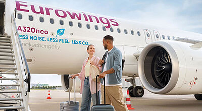 Ein junges Paar geht mit Rollkoffern eines Eurowings-Flugzeugs entlang hin zur Einstiegstreppe, um an Bord zu gehen.