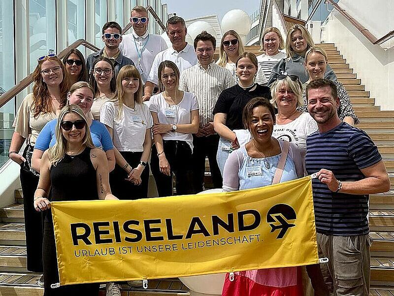 Sie stehen für die Zukunft bei Reiseland: Junge Führungskräfte der Kette bei einem Treffen im Somer 2024 auf der Aida Perla. Foto: Reiseland Eine Gruppe von etwa 20 fröhlichen Menschen steht auf einer breiten Treppe im Freien und posiert für ein Gruppenfoto. Im Vordergrund halten zwei Personen ein gelbes Banner mit der Aufschrift "REISELAND – URLAUB IST UNSERE LEIDENSCHAFT." Die Gruppe besteht aus Männern und Frauen unterschiedlichen Alters, viele tragen Namensschilder. Die Stimmung ist freundlich und entspannt, das Wetter scheint sonnig zu sein.