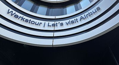 Gebäude mit gebogener Fassade und reflektierenden Fenstern. An der Außenwand steht in blauen Buchstaben: „Werkstour | Let’s visit Airbus“.