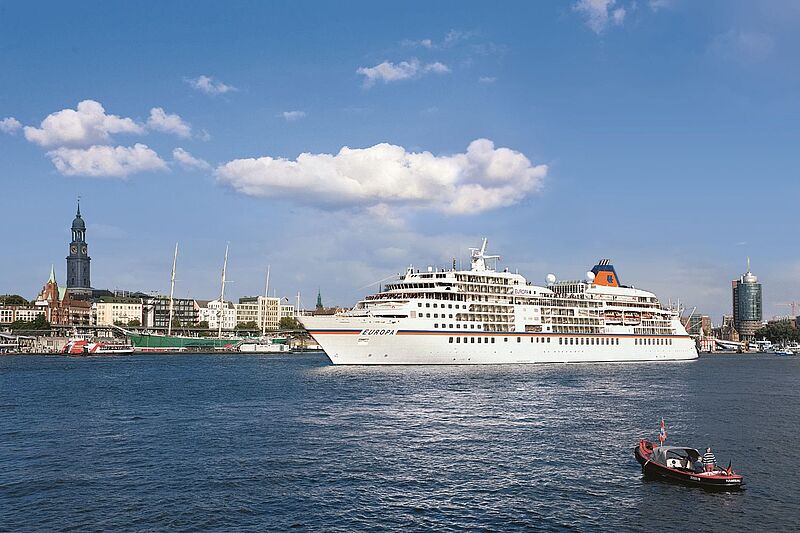 Großes Kreuzfahrtschiff „MS Europa“ fährt bei sonnigem Wetter auf einem breiten Fluss oder Hafenbecken an einer europäischen Stadt mit historischer Uferbebauung, Kirchturm und Segelschiffen vorbei; im Vordergrund ein kleines Motorboot, blauer Himmel mit wenigen Wolken.