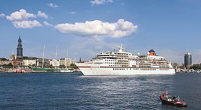 Die Europa von Hapag-Lloyd Cruises vor der Kulisse Hamburgs Großes Kreuzfahrtschiff „MS Europa“ fährt bei sonnigem Wetter auf einem breiten Fluss oder Hafenbecken an einer europäischen Stadt mit historischer Uferbebauung, Kirchturm und Segelschiffen vorbei; im Vordergrund ein kleines Motorboot, blauer Himmel mit wenigen Wolken.