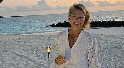 Eine lächelnde Frau steht barfuß am weißen Sandstrand bei Sonnenuntergang. Sie trägt ein luftiges, weißes Kleid mit Lochstickerei und hält ein Glas in der rechten Hand. Im Hintergrund leuchtet eine Fackel im Sand, das ruhige Meer glitzert in Blautönen, und dunkle Felsen ragen am Horizont ins Wasser. Der Himmel ist mit weichen Wolken in Pastellfarben gefärbt.
