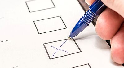 Ein weißes Blatt Papier mit Wahlkästchen. Eine Hand hält einen blauen Kugelschreiber und markiert ein Kästchen mit einem Kreuz.
