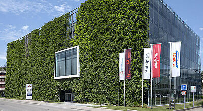 Wochen der Ungewissheit: die Zentrale von Hotelplan in der Schweiz. Foto: Hotelplan Modernes Bürogebäude mit vollständig begrünter Fassade auf der linken Seite und gläserner Front auf der rechten Seite. Vor dem Gebäude stehen vier Fahnenmasten mit den Logos von Hotelplan, Migros und weiteren Unternehmen. Der Himmel ist leicht bewölkt.