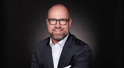 Tom Fecke ist seit Anfang Juni Global Head of Cruise bei Aviareps. Foto: Aviareps Ein Manager um die Fünfzig mit lichtem Haar und dicker Brille lächelt freundlich in die Kamera. Er trägt einen dunklen Anzug und ein weißes Oberhemd.