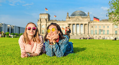 Zwei junge glückliche Frauen mit Sonnenbrille, die vor dem Bundestagsgebäude in Berlin auf den Rasen liegen