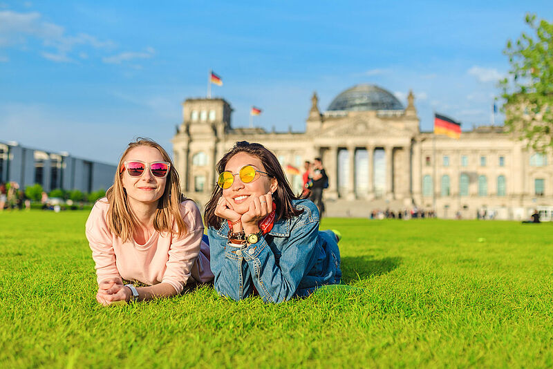 Zwei junge glückliche Frauen mit Sonnenbrille, die vor dem Bundestagsgebäude in Berlin auf den Rasen liegen