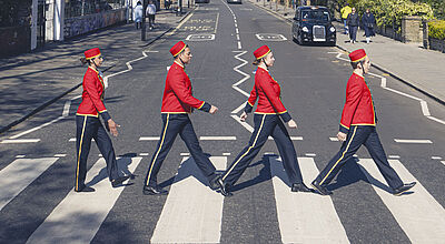 Vier Musiker in roten Uniformen überqueren im Gleichschritt einen Zebrastreifen auf der Abbey Road in London, ähnlich dem berühmten Album-Cover.