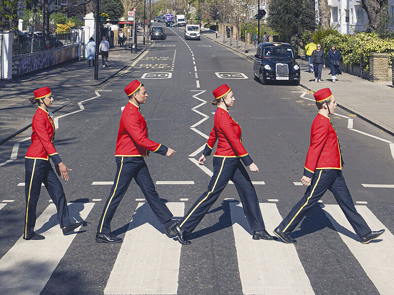 Vier Musiker in roten Uniformen überqueren im Gleichschritt einen Zebrastreifen auf der Abbey Road in London, ähnlich dem berühmten Album-Cover.