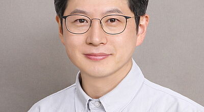 Porträt eines lächelnden asiatischen Mannes mit Brille und weißem Hemd vor grauem Hintergrund.