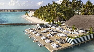 Luftaufnahme eines luxuriösen Malediven-Resorts mit Overwater-Bungalows, Holzterrasse mit weißen Liegen und Sonnenschirmen über türkisblauem Wasser und Palmen am Strand