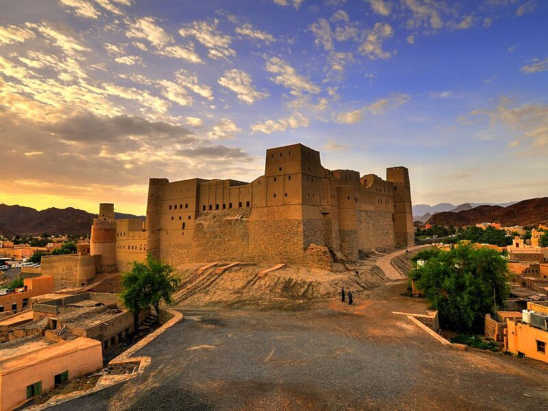 Eine große, sandfarbene Festung aus Lehm und Stein erhebt sich auf einem Hügel in einer Wüstenlandschaft bei Sonnenuntergang. Der Himmel ist teils bewölkt und warm gefärbt. Rund um die Festung liegen traditionelle Häuser und grüne Bäume; zwei Menschen in dunkler Kleidung gehen einen Pfad entlang vor der gewaltigen Burganlage.