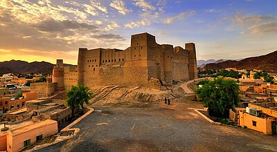 Eine große, sandfarbene Festung aus Lehm und Stein erhebt sich auf einem Hügel in einer Wüstenlandschaft bei Sonnenuntergang. Der Himmel ist teils bewölkt und warm gefärbt. Rund um die Festung liegen traditionelle Häuser und grüne Bäume; zwei Menschen in dunkler Kleidung gehen einen Pfad entlang vor der gewaltigen Burganlage.