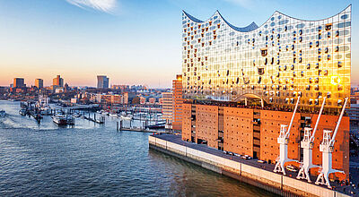 Elbphilharmonie im Sonnenuntergang am Hamburger Hafen mit glitzernder Glasfassade, Hafenkränen und Bootsliegeplätzen im Vordergrund