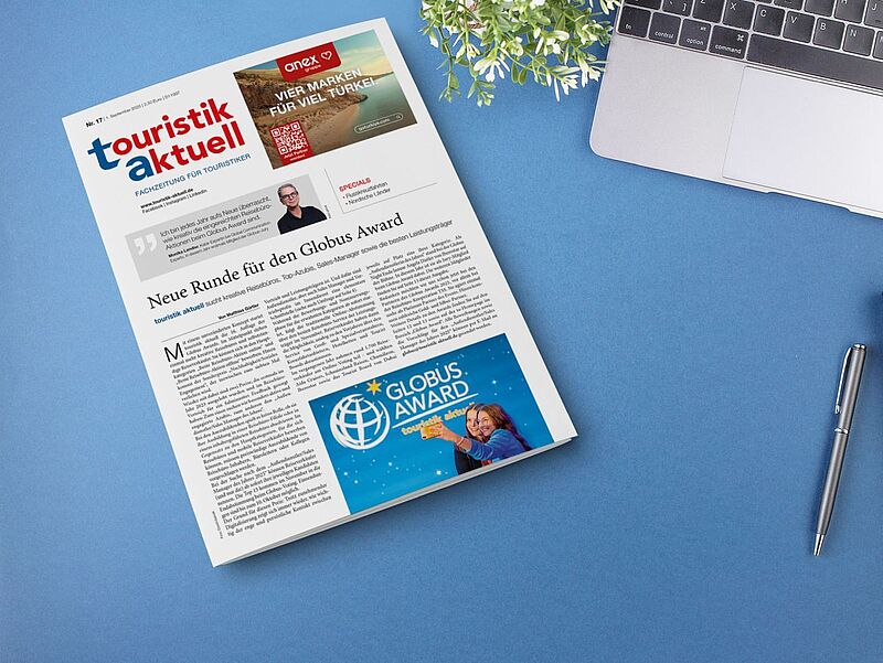 touristik aktuell kann als gedruckte Ausgabe sowie als E-Paper gelesen werden. Fotonachweis: Roman-Valiev/iStock Auf einem blauen Schreibtisch liegt eine Ausgabe der Fachzeitung touristik aktuell mit dem Titelthema ‚Neue Runde für den Globus Award‘. Neben der Zeitung befinden sich ein aufgeklappter Laptop, ein schwarzes Notizbuch mit Stift und eine kleine grüne Pflanze.“