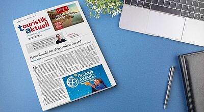 Auf einem blauen Schreibtisch liegt eine Ausgabe der Fachzeitung touristik aktuell mit dem Titelthema ‚Neue Runde für den Globus Award‘. Neben der Zeitung befinden sich ein aufgeklappter Laptop, ein schwarzes Notizbuch mit Stift und eine kleine grüne Pflanze.“