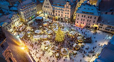 Luftaufnahme des verschneiten Weihnachtsmarkts in Tallinn mit beleuchtetem Tannenbaum und Menschenmenge am Abend.