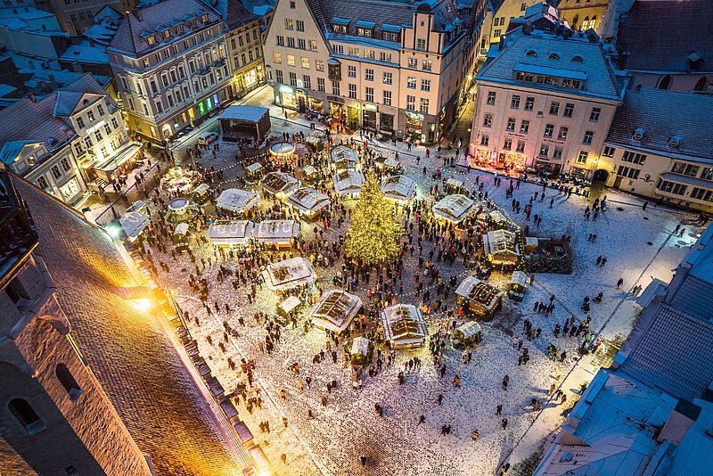 Luftaufnahme des verschneiten Weihnachtsmarkts in Tallinn mit beleuchtetem Tannenbaum und Menschenmenge am Abend.
