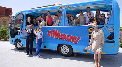 Mit dem Shuttle-Bus ging es durch die Pickalbatros-Hotels Villaggio Resort und Vita Resort In einem blauen Shuttle-Bus sitzen mehrere Personen und schauen durch die geöffneten Fenster. Vor dem Bus stehen ein Mann und zwei Frauen, die ein blaues Banner in den Händen halten, auf dem Alltours zu lesen ist