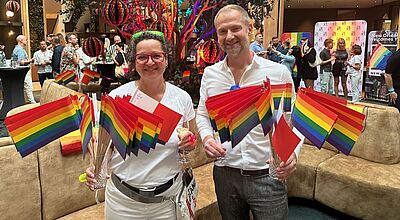 Zwei Personen in weißer Kleidung stehen lächelnd in einem farbenfroh dekorierten Innenraum, halten zahlreiche Regenbogenfahnen in den Händen und ein Glas Sekt. Im Hintergrund feiern viele Menschen eine Pride-Veranstaltung mit bunten Dekorationen, Regenbogenfarben und einem beleuchteten Baum in der Mitte. Banner im Hintergrund tragen Aufschriften wie „New Orleans welcomes you“.