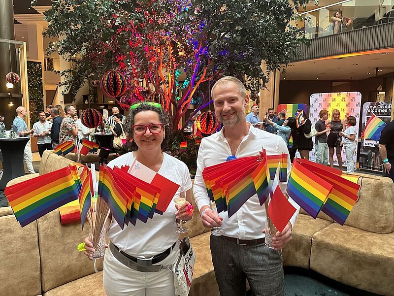 Zwei Personen in weißer Kleidung stehen lächelnd in einem farbenfroh dekorierten Innenraum, halten zahlreiche Regenbogenfahnen in den Händen und ein Glas Sekt. Im Hintergrund feiern viele Menschen eine Pride-Veranstaltung mit bunten Dekorationen, Regenbogenfarben und einem beleuchteten Baum in der Mitte. Banner im Hintergrund tragen Aufschriften wie „New Orleans welcomes you“.