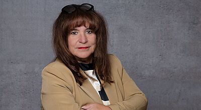 Porträt einer Frau mit braunen Haaren und Pony, die eine beige Jacke trägt. Sie steht mit verschränkten Armen vor einem grauen Hintergrund und trägt eine Brille auf dem Kopf.