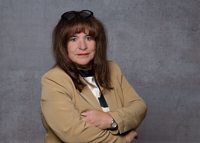 Marija Linnhoff ist bislang die einzige Kandidatin für das Amt der DRV-Präsidentin. Foto: VUSR Porträt einer Frau mit braunen Haaren und Pony, die eine beige Jacke trägt. Sie steht mit verschränkten Armen vor einem grauen Hintergrund und trägt eine Brille auf dem Kopf.