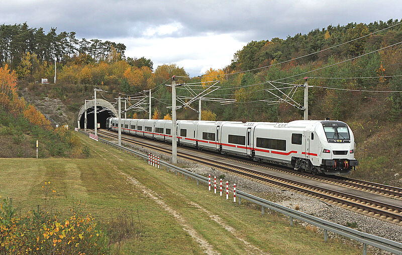 Ein weißer ICE-Zug des neuen Modells ICE L fährt in einen Tunnel auf elektrifizierter Bahnstrecke, umgeben von herbstlicher Vegetation und bewölktem Himmel.