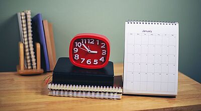 Ein roter Wecker, der 10:52 Uhr anzeigt, steht auf einem Stapel Notizbücher neben einem Tischkalender für den Monat Januar. Im Hintergrund befinden sich weitere aufrecht stehende Notizbücher in einem Holzhalter vor einer grünen Wand.