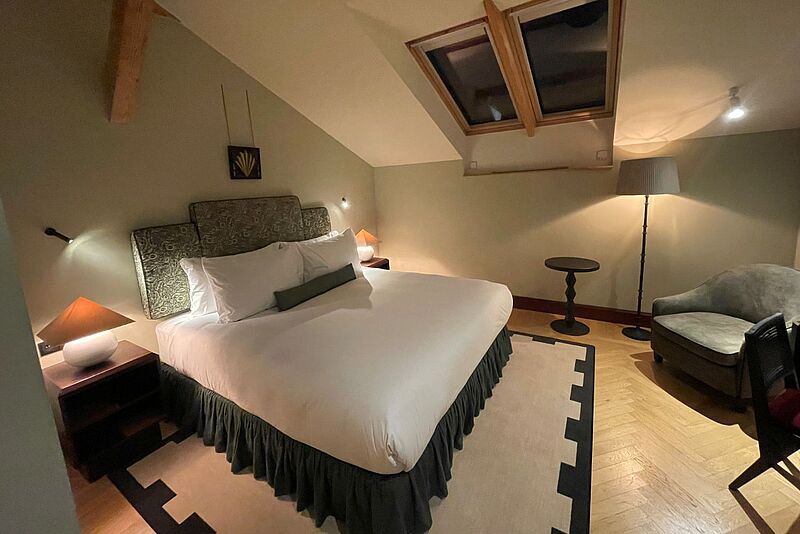 Die Zimmer im Sir Prague sind von der Gestaltung her an den Kubismus angelehnt. Bild von einem indirekt beleuchteten Hotelzimmer mit Dachschräge, in dem ein Doppelbett mit Nachtschränken und ein Sessel stehen.
