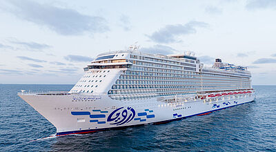 Die Aroya wurde auf der Meyer Werft in Deutschland gebaut – ursprünglich als World Dream für den pleitegegangenen chinesischen Anbieter Dream Cruises. Foto: Aroya Cruises Ein neues saudisches Kreuzfahrtschiff mit weißem Raumpf und Aufbau fährt auf blauem Meer. Am Rumpf sind aufgehängte rot-weiße Rettungsboote und aufgemalte blaue Rechtecke zu sehen.