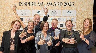 Luxusurlaub, Kiticon, Silvio Rebmann, Cuba Eine Gruppe von sieben fröhlichen Personen posiert vor einer goldenen Fotowand mit der Aufschrift „INSPIRE ME AWARD 2025“. Alle halten gläserne Trophäen in den Händen und lächeln in die Kamera. Im Hintergrund sind Logos verschiedener Sponsoren zu sehen.