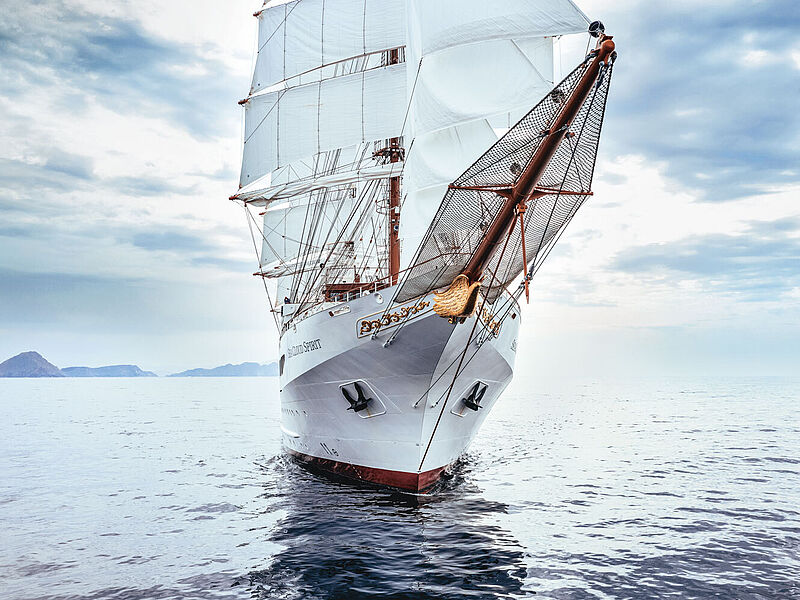 Moderner Windjammer: Die Sea Cloud Spirit verfügt unter anderem über Balkonkabinen und ein Bistro als Alternative zum Fine-Dining-Restaurant. Foto: Sea Cloud Cruises Frontalansicht eines schneeweißen Luxus-Segelkreuzfahrtschiffes, das auf blauem Meer unterwegs ist.