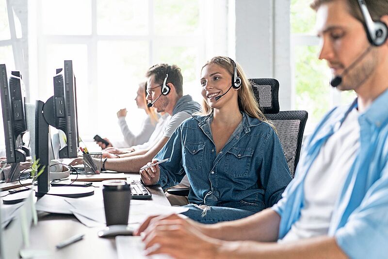 Für viele Reisebüro-Mitarbeiter ist eine gute und persönliche Betreuung seitens der Leistungsträger sehr wichtig Mehrere junge Mitarbeiterinnen und Mitarbeiter in einem modernen Callcenter arbeiten mit Headsets an Computern. Eine Frau im Vordergrund lächelt während des Gesprächs.