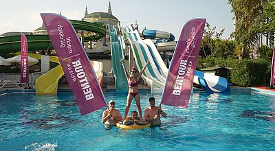 Die Teilnehmer der Bentour Reisen Aquapark Championship 2025 hatten viel Spaß Vier Personen stehen in einem Pool vor großen Wasserrutschen in Grün, Weiß und Blau. Zwei Männer halten aufblasbare rosa Bentour-Reisen-Werbebanner, ein Mann liegt entspannt auf einem gelben Schwimmring, und eine Frau in einem pinken Bikini steht mit erhobenen Armen auf dem Ring. Im Hintergrund sind grüne Wasserrutschen, Sonnenschirme und Vegetation zu sehen. Über der Szenerie ragen die Türme eines Gebäudes im orientalischen Stil hervor. Die Stimmung wirkt fröhlich und sommerlich.