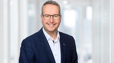 Stefan Kirchner, Vertriebsmanager bei der Allianz, steht lächelnd vor hellem Büro-Hintergrund. Er ist trägt ein helles Hemd, ein Sakko und eine Brille.