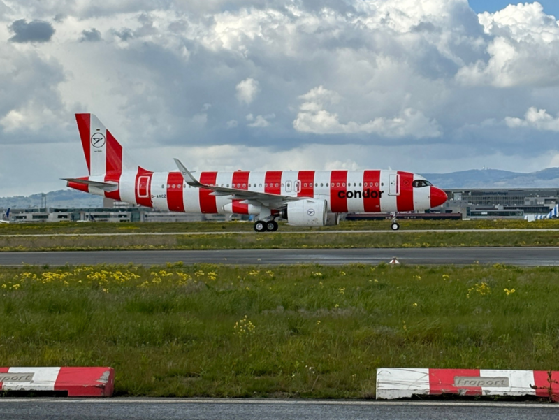 Ein Airbus A320neo der Fluggesellschaft Condor mit rot-weiß gestreifter Lackierung rollt über das Rollfeld des Frankfurter Flughafens bei bewölktem Himmel. Im Hintergrund sind Flughafengebäude und das Bergpanorama des Taunus zu sehen. Im Vordergrund befindet sich eine mit "Fraport" gekennzeichnete rot-weiße Begrenzung.