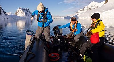 Auch bei Antarktis-Reisen mit HX Hurtigruten Expeditions können Kunden sparen Zwei Personen in blauen Jacken und eine in gelber Jacke sitzt auf einem Zodiac in der Antarktis, im Hintergrund sieht man Eisberge