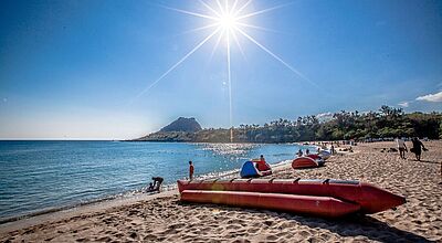 Ein sonniger Tag am Nanwan Beach in der taiwanischen Pingtung-Region. Der Strand ist belebt mit Menschen, die im Sand entspannen oder im Wasser spielen. Mehrere rote Schlauchboote liegen auf dem Sand, während im Hintergrund ein bewaldeter Hügel die Szenerie abrundet. Die Sonne steht hoch am Himmel und erzeugt starke Lichtstrahlen und Reflexionen auf dem Meer.