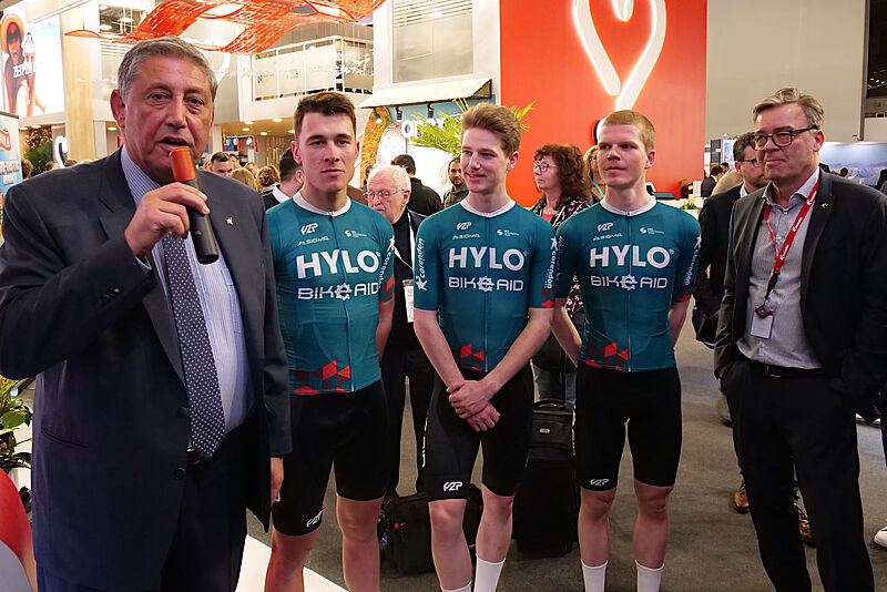 Zwei Manager stehen zusammen mit einem Rennrad-Team auf dem Messestand der Airline Corendon auf der Reisemesse ITB in Berlin.