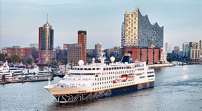 Das kleine Kreuzfahrtschiff Hamburg mit blauweißem Rumpf verlässt den Hafen der gleichnamigen Hansestadt. Im Hintergrund thront vor blauem Himmel die Elbphilharmonie.